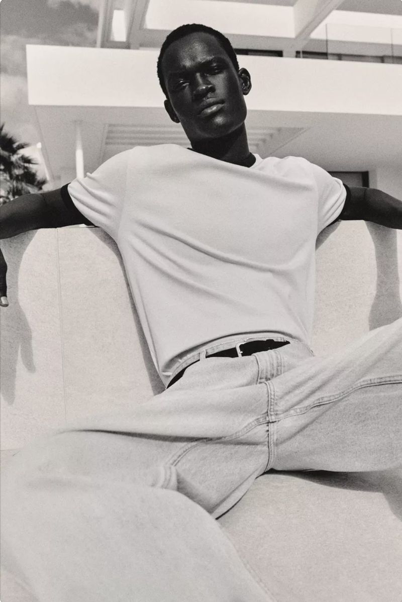 Monochrome portrait of a dark-skinned adult male model sitting in casual wear outdoors with architecture and palm trees visible.