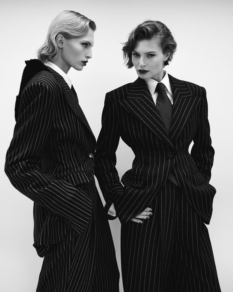 Two women in pinstripe suits pose in a studio, showcasing classic tailoring and monochrome style with short, styled hair and dark lipstick.
