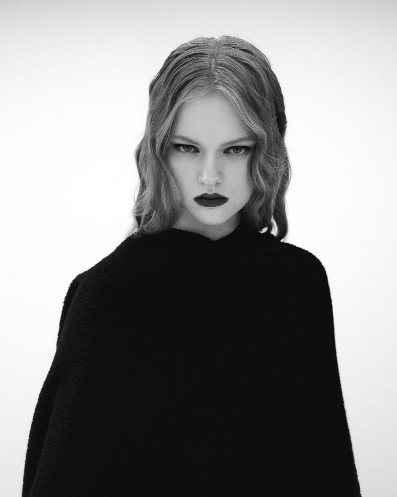 Monochrome portrait of a young woman with wavy blond hair and dark lipstick, wearing a dark fabric against a plain white background.