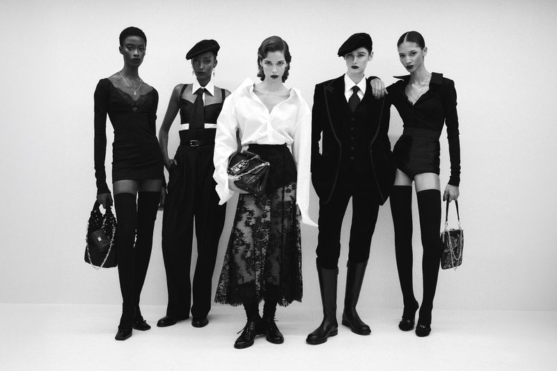 Five fashion models pose in a monochromatic photo studio wearing chic black and white apparel, including berets and thigh-high socks.
