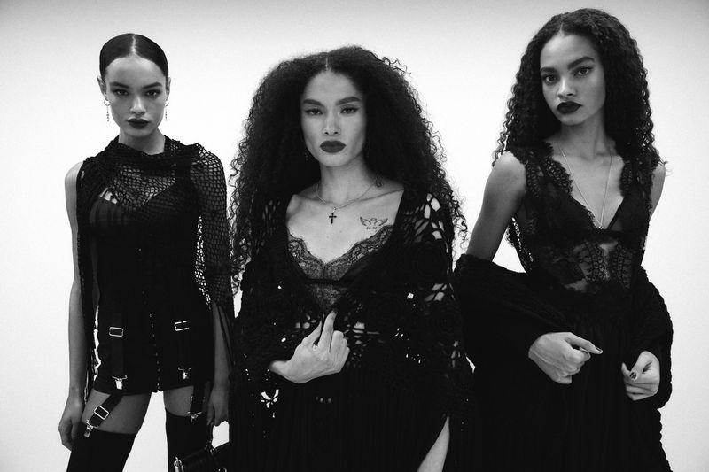Three adult women pose in a studio, showcasing black lace tops, dark lipstick, and curly hair against a plain white background for fashion.