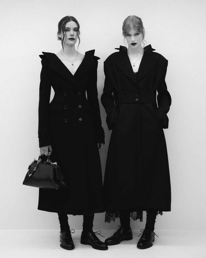 Two fashion models in black dresses and coats pose in a monochrome studio setting, one holding a black purse, showcasing high-fashion style.