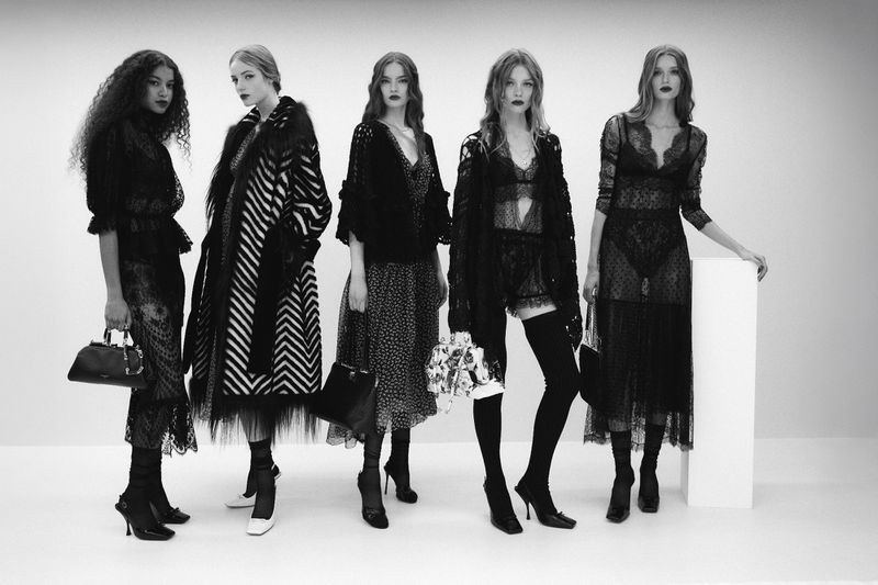 Five models in black lace dresses and patterned coats standing in a row against a plain white studio background.