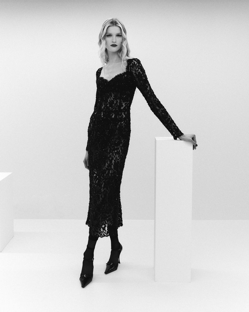 Blonde fashion model poses in long-sleeved black lace dress, black pointed heels, and red lipstick in front of white cubes.