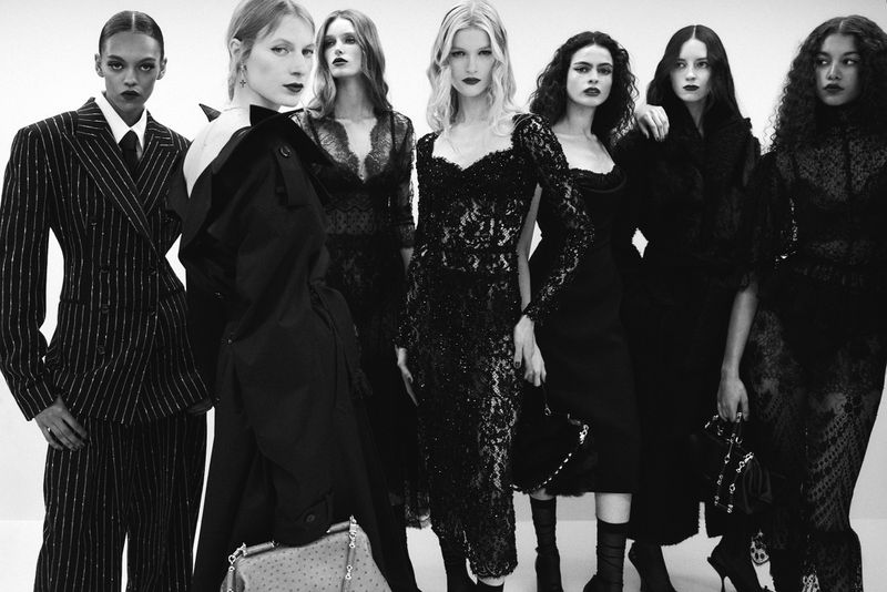 Seven adult women in various black designer outfits pose in a stark white photo studio, monochromatic, elegant and moody.