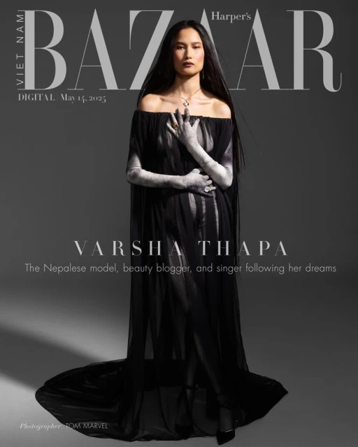Varsha Thapa posing in Harper's Bazaar cover wearing a flowing black dress and jewelry with artistic hand makeup against a gray background.