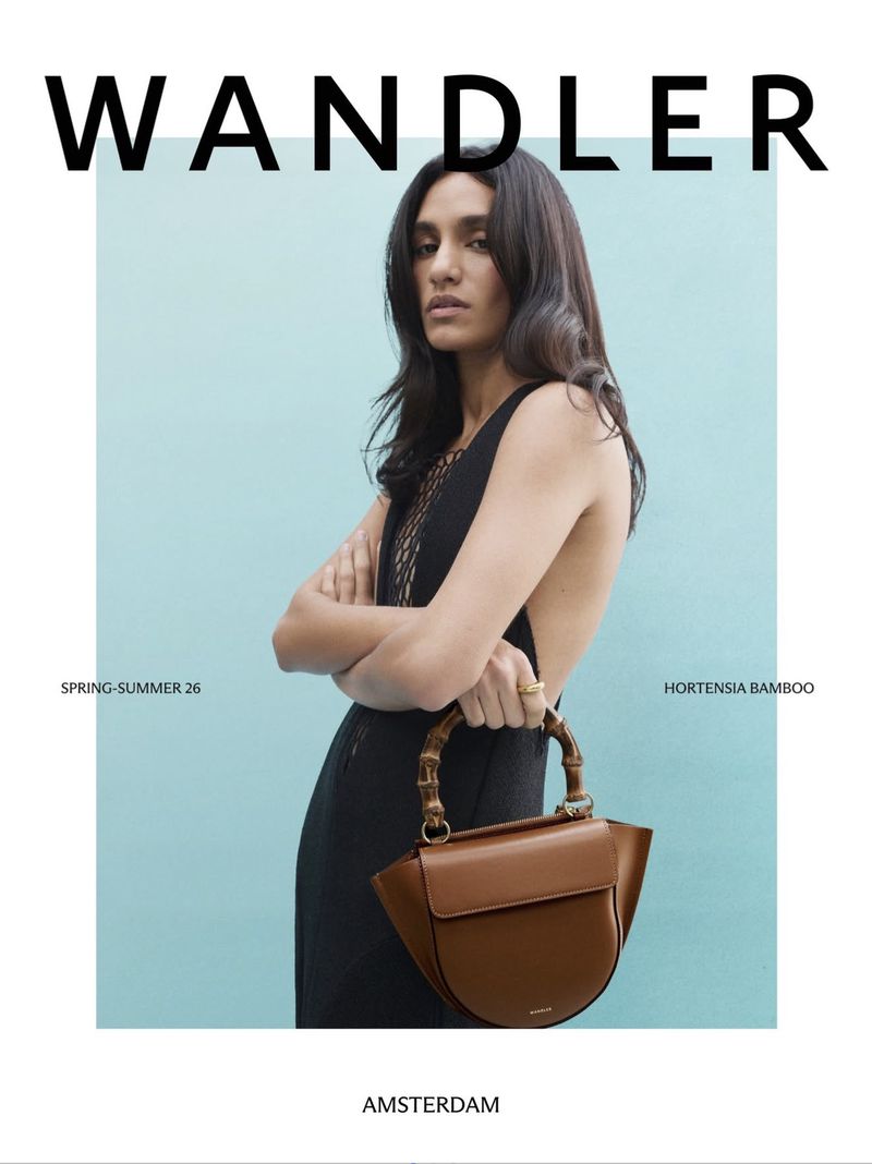 Fashion editorial shot of a woman with long dark hair holding a tan leather handbag with bamboo handles in front of an aqua background.