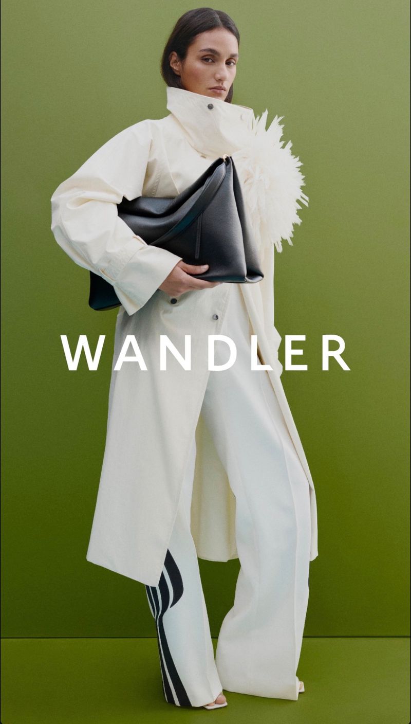 A model holding a large black leather handbag against an olive green background, wearing a cream trench coat with feather embellishment.