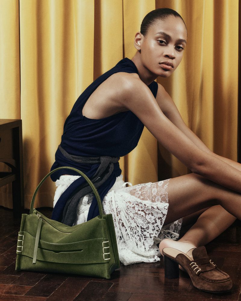 A person sitting on a wooden floor with a green handbag, wearing a dark top and light skirt, with a yellow curtain backdrop.