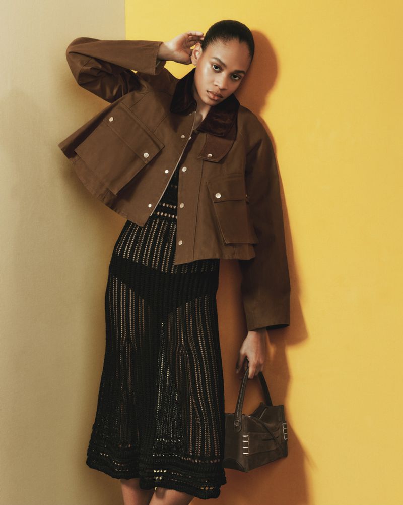 A woman in a brown jacket and patterned dress poses against a yellow and beige background, holding a structured brown handbag.