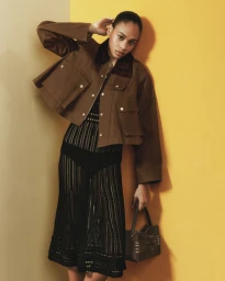 Model wearing a brown jacket and black patterned dress posing against a yellow and cream studio wall for J.W. Anderson