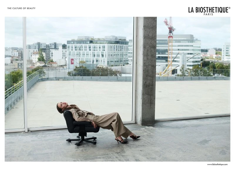 Woman in suit reclines in office chair by expansive window overlooking cityscape. La Biosthetique Paris advertisement.