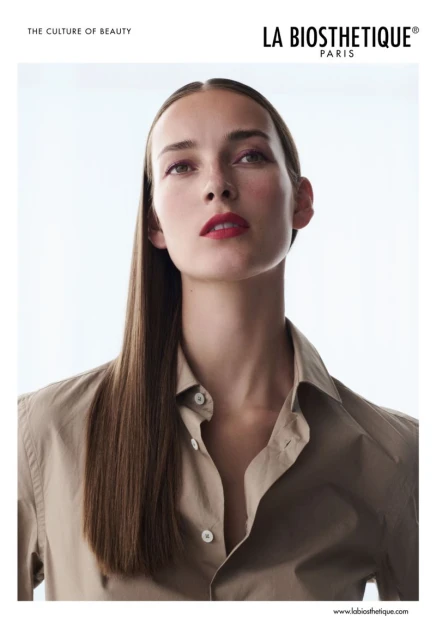 Beauty portrait of a woman with straight brown hair, wearing a tan button-down shirt, against a bright white background. Cosmetics advertisement.