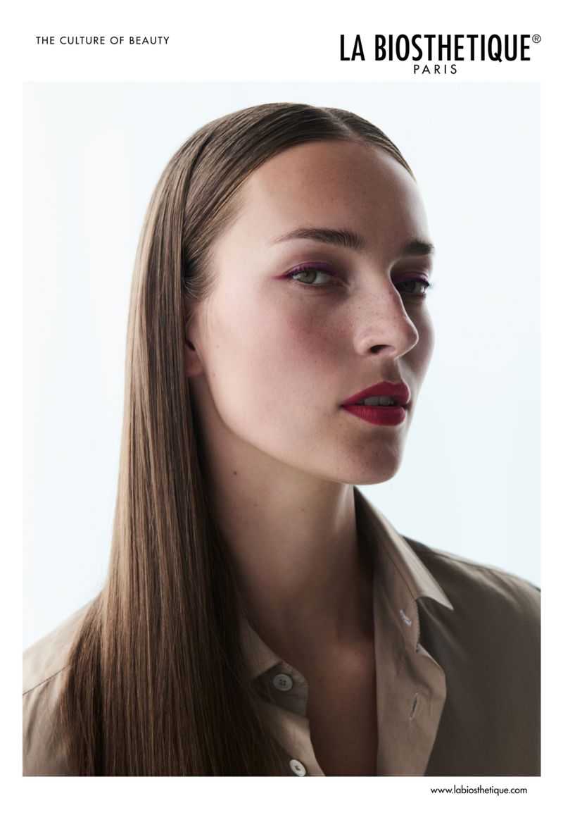 Beauty shot of a young adult woman with long, straight brown hair, wearing a beige button-up shirt, in a brightly lit indoor studio.