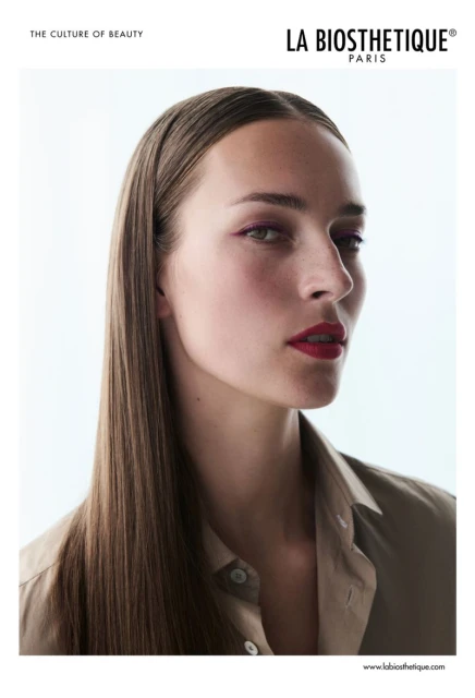 Beauty shot of a young adult woman with long, straight brown hair, wearing a beige button-up shirt, in a brightly lit indoor studio.