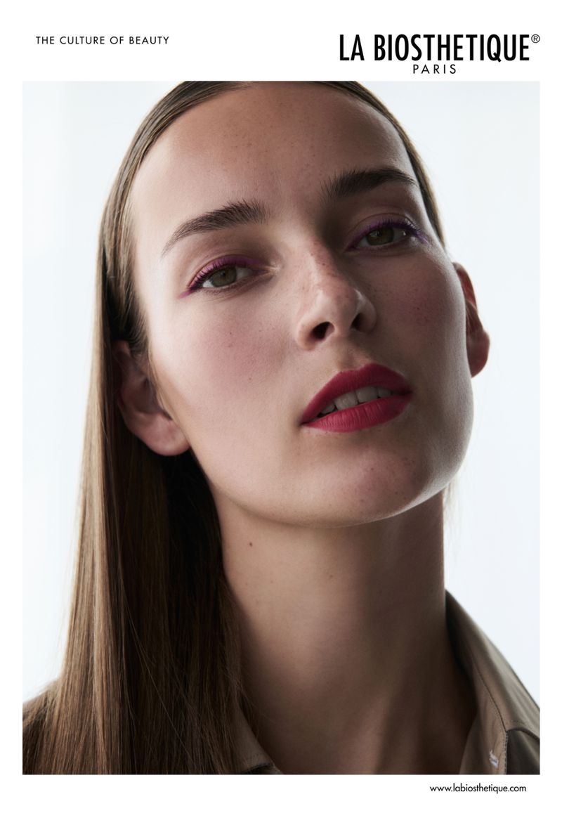 Close-up portrait of a woman with red lipstick and straight brunette hair, wearing a neutral shirt, in a beauty advertisement.