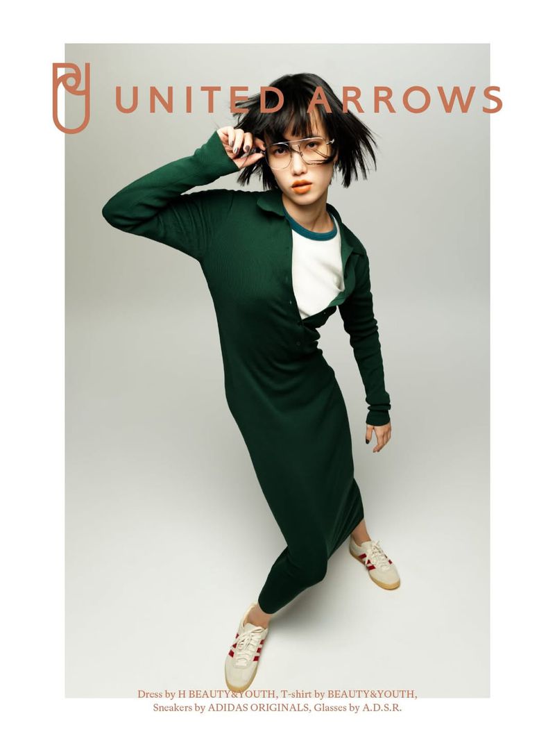 Fashion shot of a woman in a green dress and glasses, posing on a grey background with ADIDAS sneakers, for United Arrows brand.