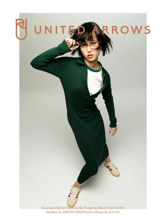 Fashion shot of a woman in a green dress and glasses, posing on a grey background with ADIDAS sneakers, for United Arrows brand.