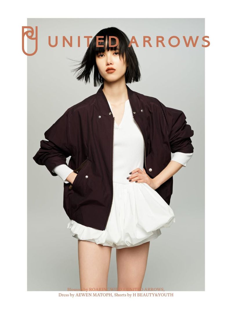 A woman poses indoors wearing a dark brown bomber jacket, white top, and white bloomers; fashion shot for United Arrows brand.
