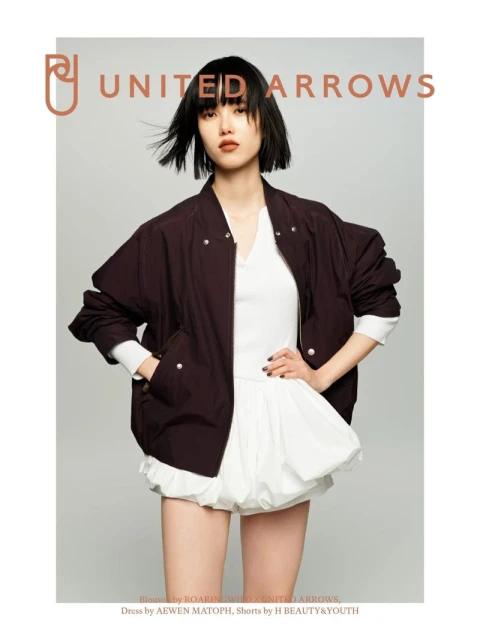 A woman poses indoors wearing a dark brown bomber jacket, white top, and white bloomers; fashion shot for United Arrows brand.
