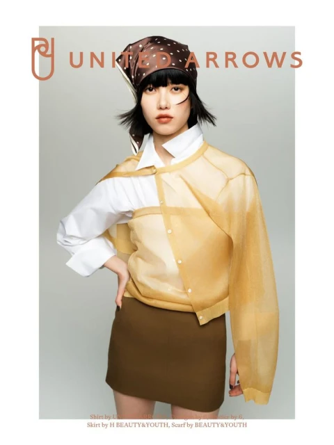 Adult female fashion model poses indoors in a light brown mini skirt, white collared shirt, and gold sheer button-down cardigan.