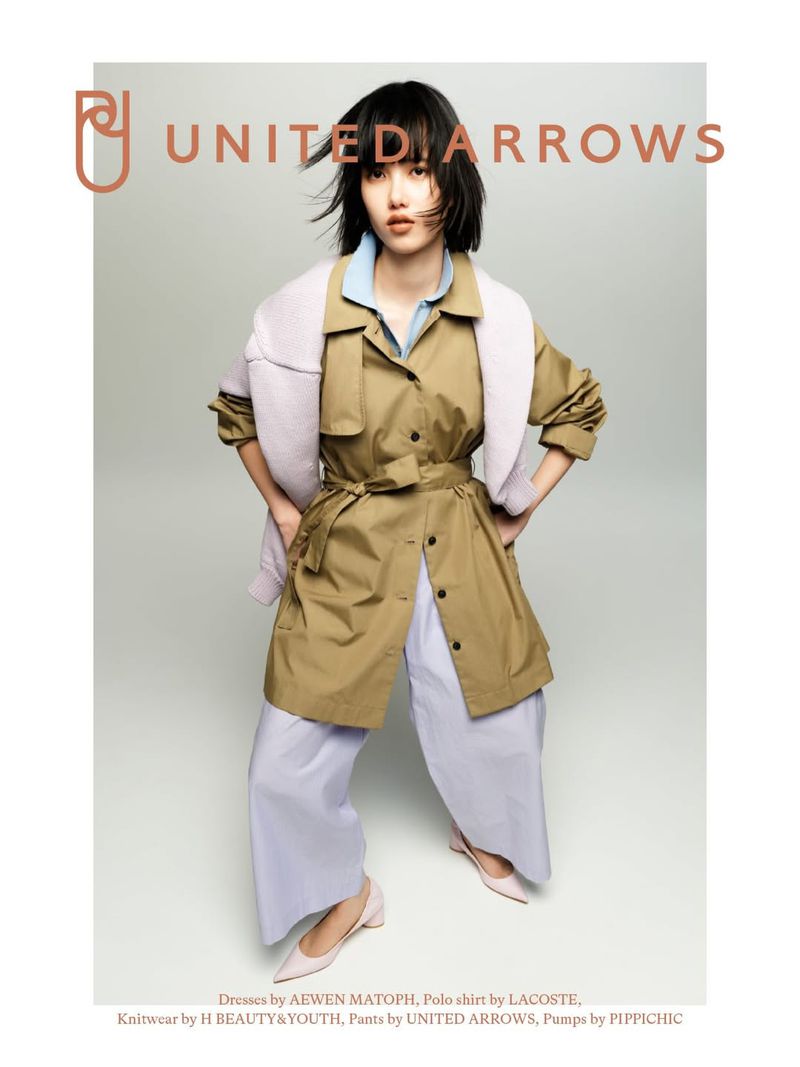 A young woman poses in a light brown trench coat, pale blue blouse and lavender pants. Soft light and muted colors create a calm, modern fashion aesthetic.