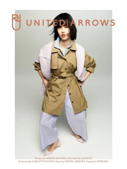 A young woman poses in a light brown trench coat, pale blue blouse and lavender pants. Soft light and muted colors create a calm, modern fashion aesthetic.
