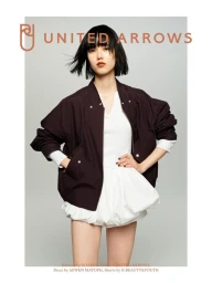 Model in dark brown bomber jacket and white shorts posing against a grey seamless studio backdrop with brand text overlay.