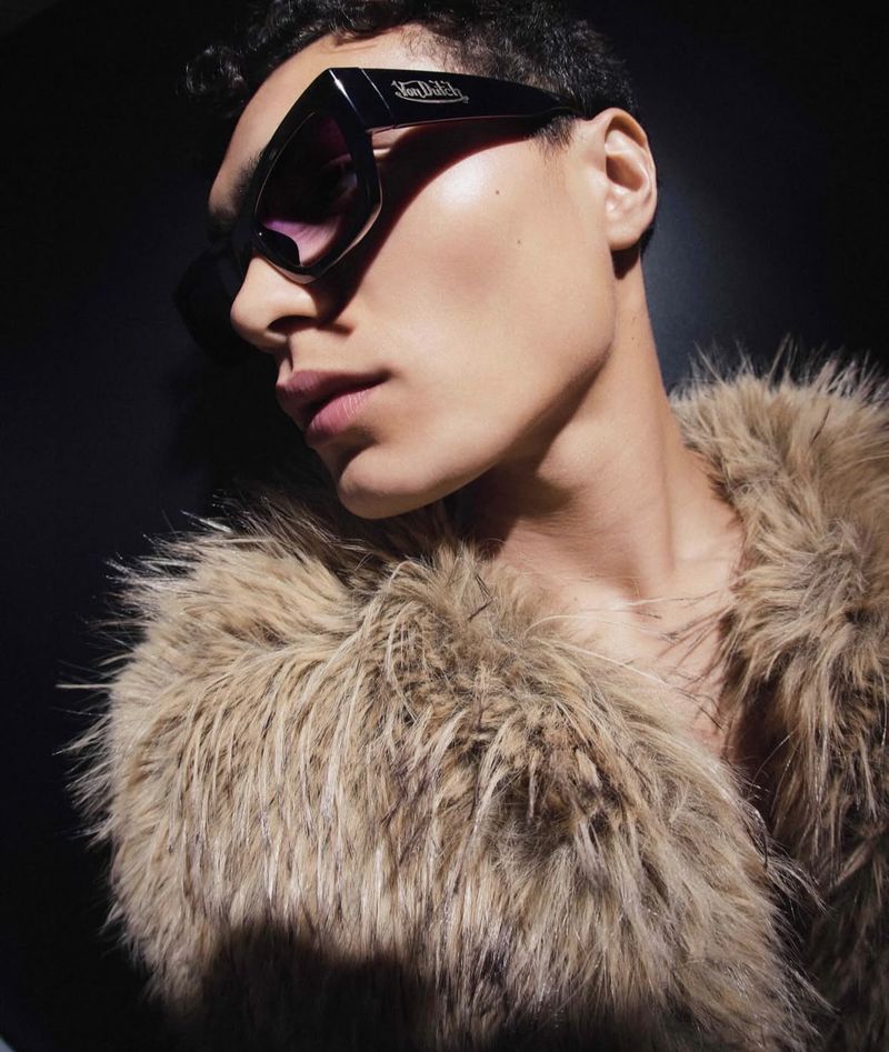 Close-up of a stylish adult in sunglasses and a faux fur coat, looking to the side against a dark background with dramatic lighting.