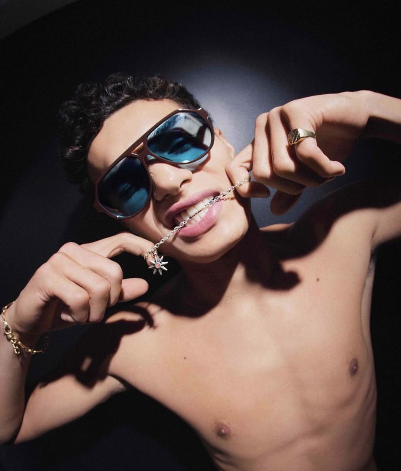 A young adult male with sunglasses is biting a silver necklace with a star pendant in a dark, high-contrast studio portrait.