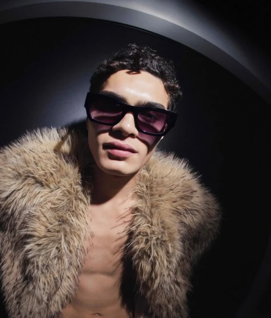Stylish young adult man in sunglasses wearing a luxurious faux fur coat against a dark backdrop. Fashion and portrait photography.