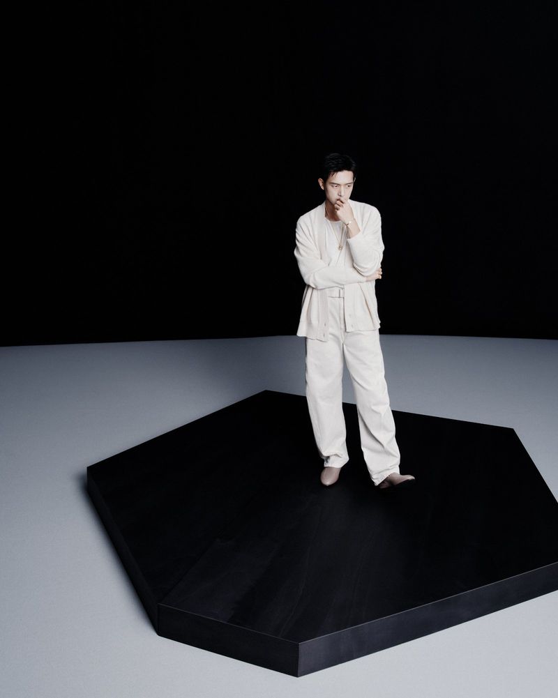 An adult male model in light-colored clothing poses pensively on a black platform against a dark backdrop in a full shot.