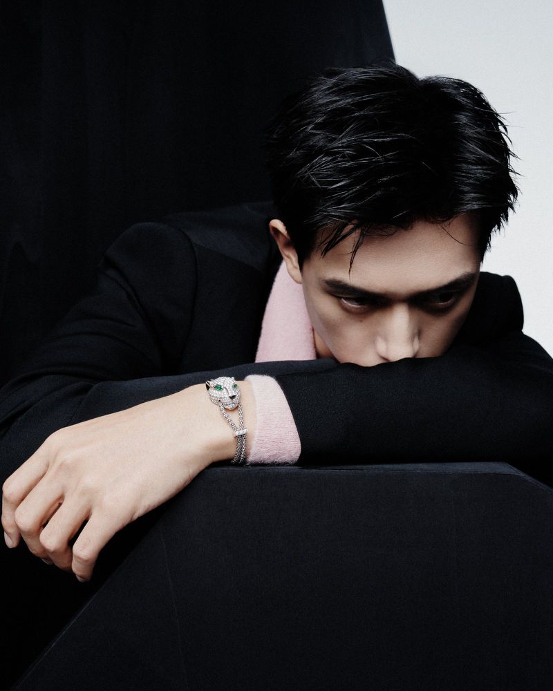 Asian man in a black suit with silver panther bracelet leans on black chair in a studio setting. Close-up portrait with formal fashion.