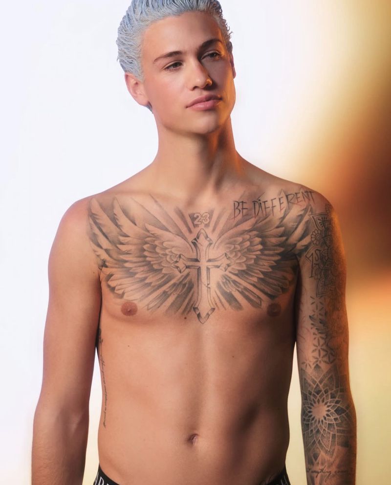 Portrait of a young adult man with silver dyed hair and torso tattoos, posing shirtless indoors against a bright, gradient background.
