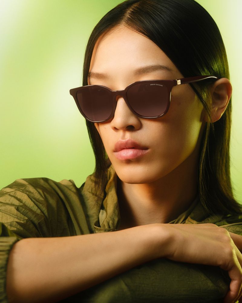Woman with dark hair models brown-framed sunglasses in a studio setting with soft, diffused green lighting and natural makeup.