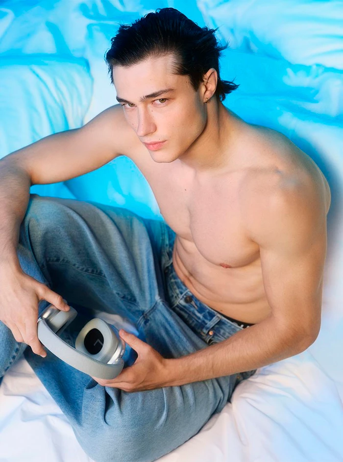 Shirtless young man sitting on textured white bedding holding grey headphones in blue ambient light