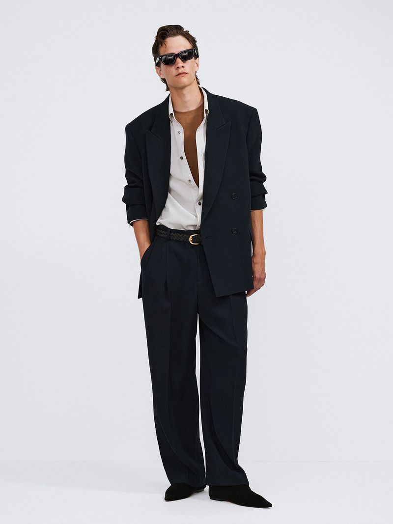 Male model poses in a double-breasted blazer, wide-legged pants, and sunglasses against a white background in a fashion editorial.