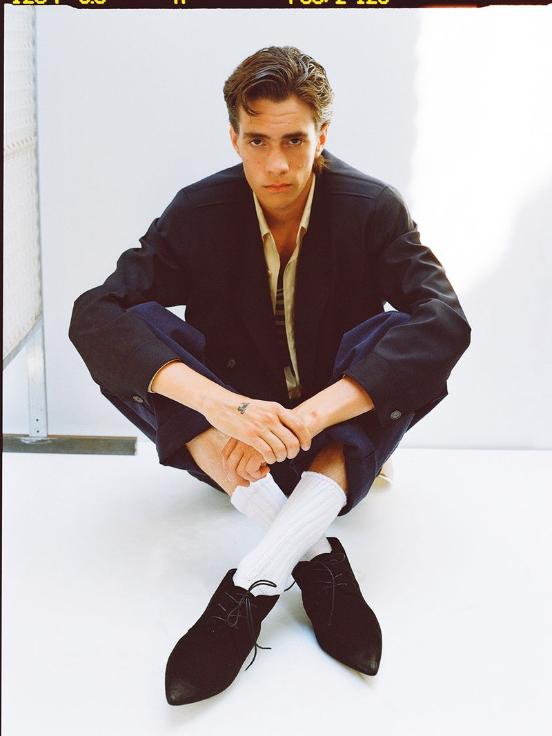 Model seated on white studio floor wearing a dark jacket, blue trousers, white socks, and black shoes