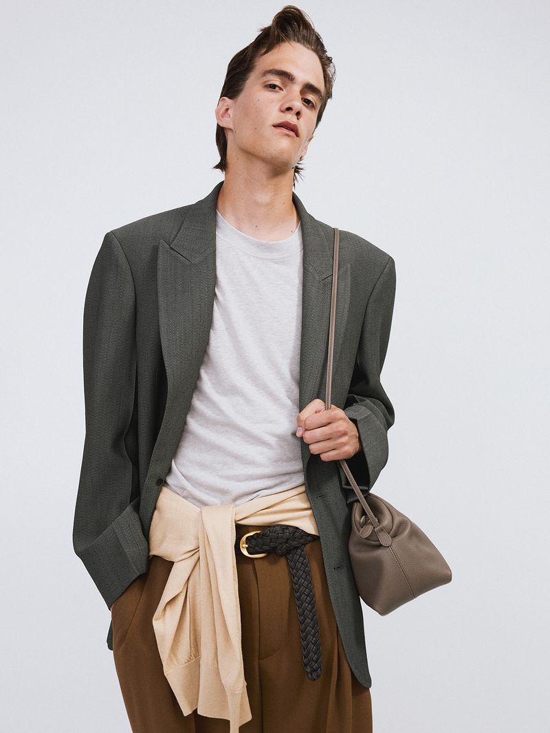 Male model in a grey blazer, white t-shirt, and brown pants, posing in a studio with a taupe-colored purse slung over his shoulder.