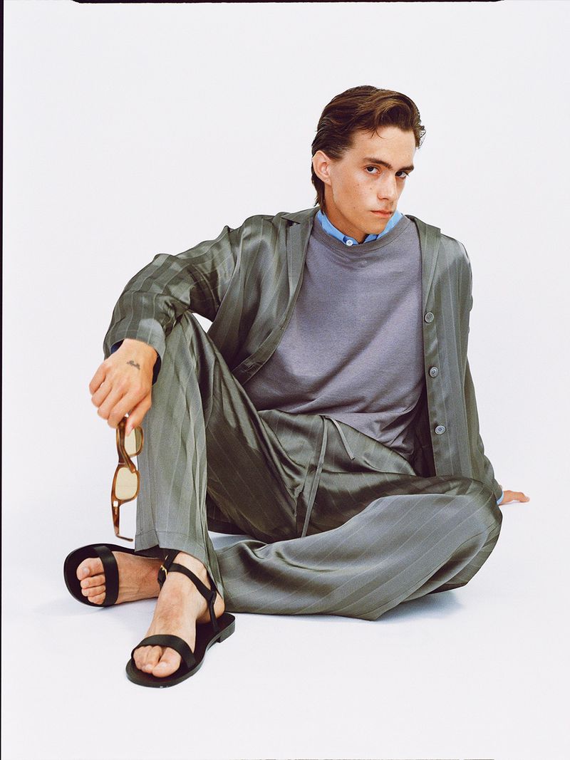Stylish young man in a relaxed pose wearing a gray striped satin suit and black sandals, holding sunglasses, in a bright photo studio.