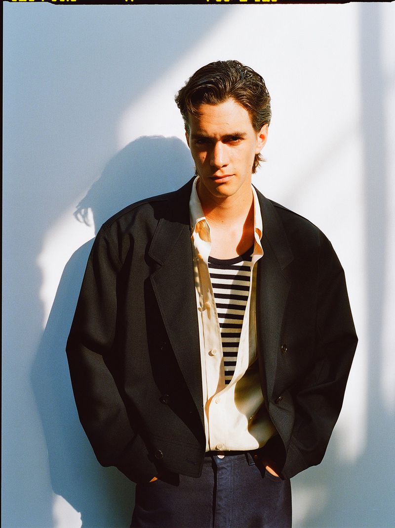 Portrait of a young adult man posing in a black blazer, striped shirt, and dark jeans against a white wall with shadow.
