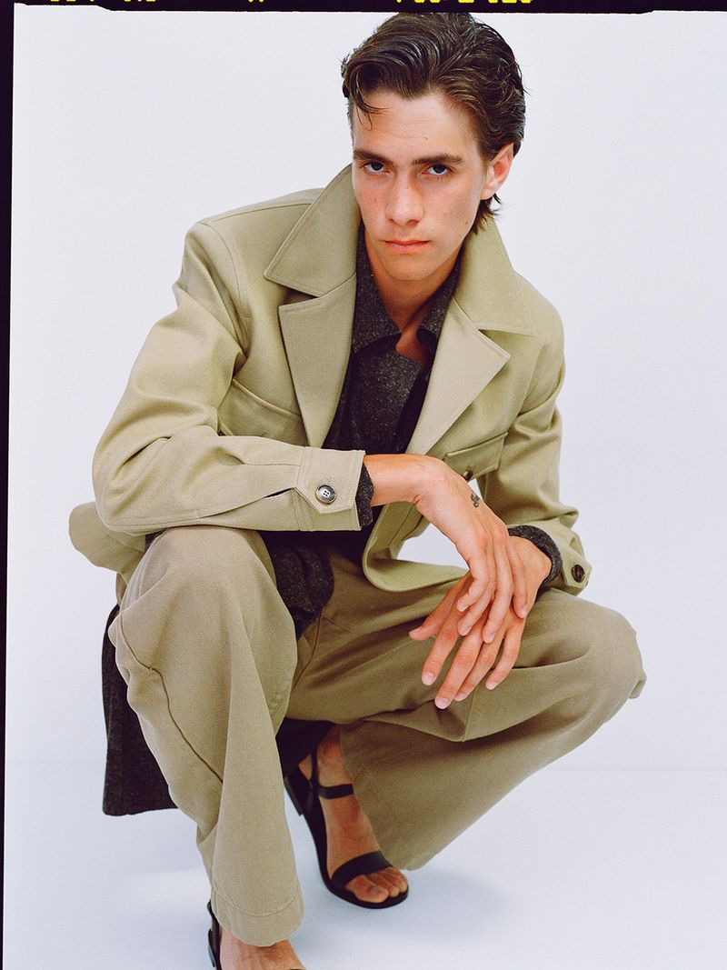 A young adult male with dark hair poses in a beige jacket, pants, and black sandals against a white backdrop in a studio shot.