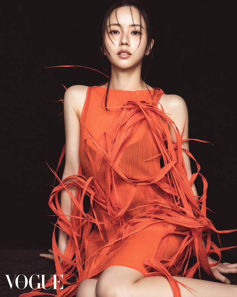 Portrait of an Asian woman in an orange dress with fringed detail, posing indoors against a dark background for a fashion editorial.