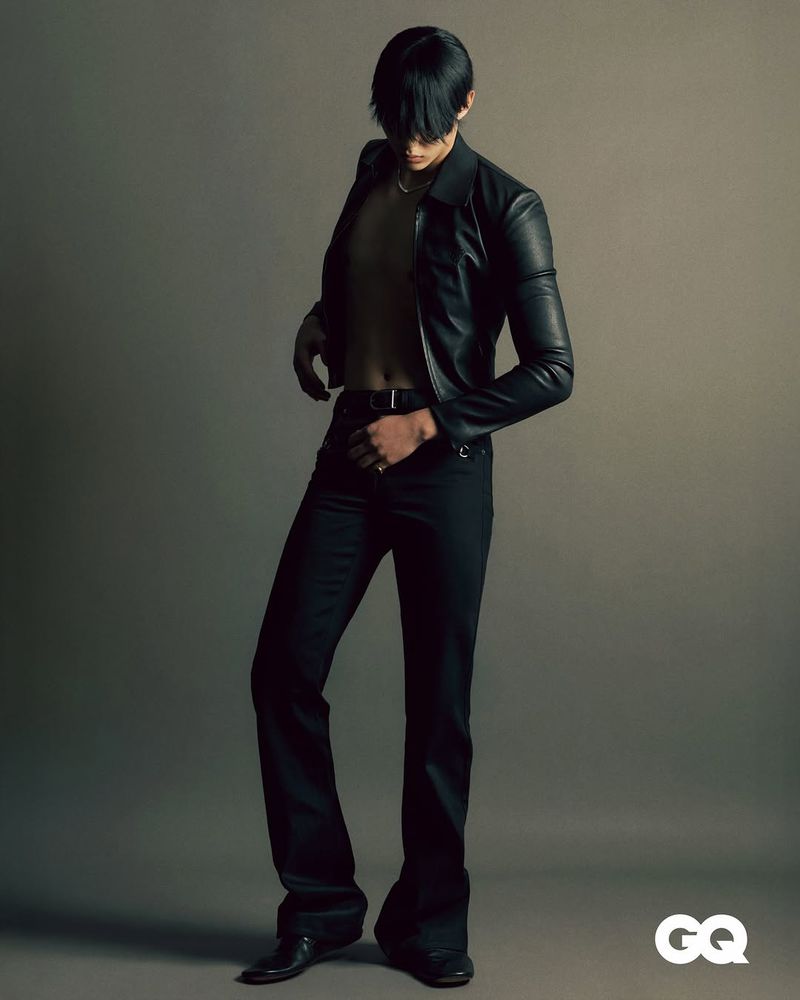 Fashionable man in leather jacket and flared pants poses in a studio, showcasing a blend of classic and contemporary style.