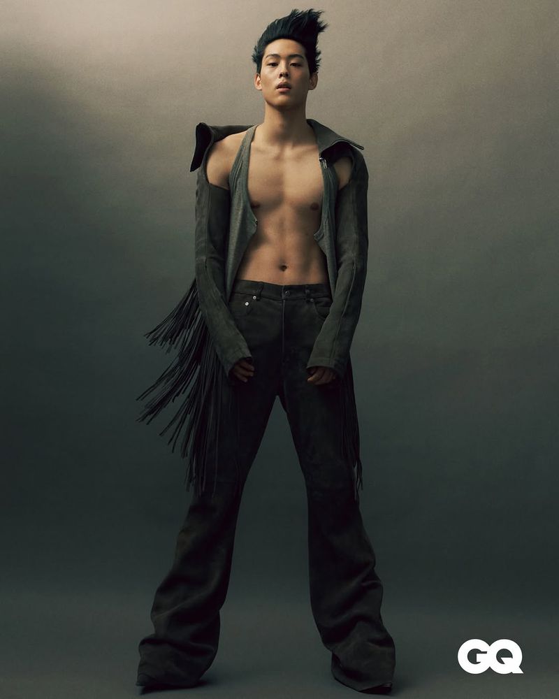 Full-length portrait of an adult male model posing in a jacket with fringe and wide-leg pants against a gradient grey background.