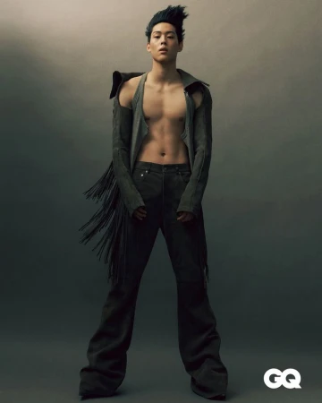 Full-length portrait of an adult male model posing in a jacket with fringe and wide-leg pants against a gradient grey background.