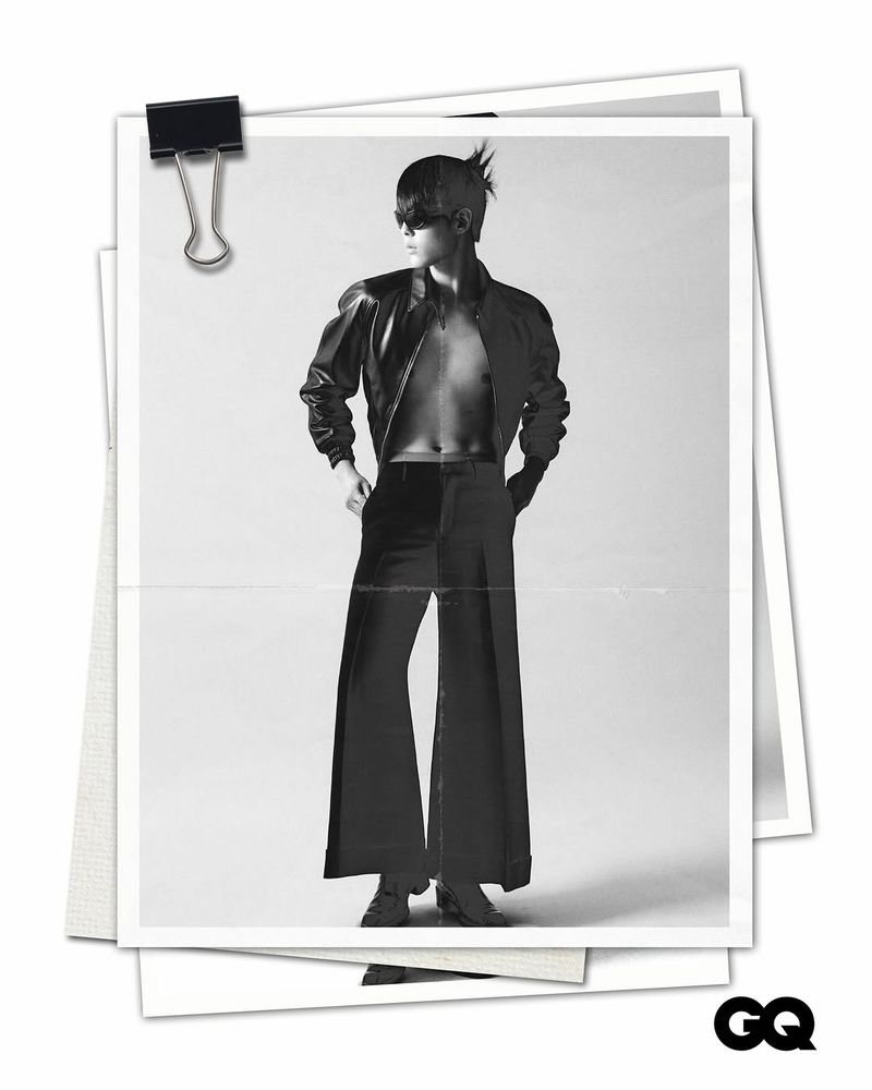 Black and white photograph on paper of a person with spiky hair wearing a leather jacket, dress pants, and heeled shoes against light backdrop.