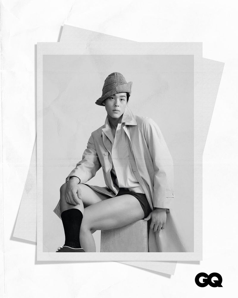 Monochrome portrait of a seated Asian man in shorts, long socks, bucket hat, and trench coat, photographed on a set of printed papers.