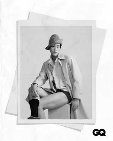 Monochrome portrait of a seated Asian man in shorts, long socks, bucket hat, and trench coat, photographed on a set of printed papers.