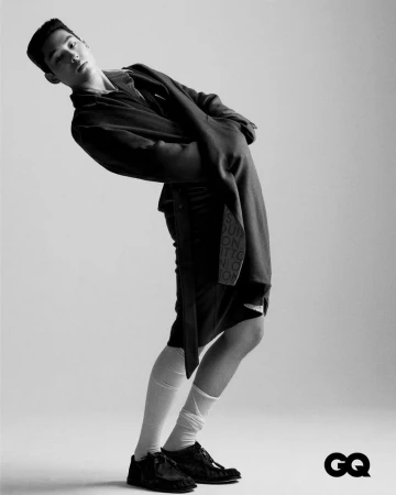Monochromatic fashion portrait of a male model wearing a dark jacket, shorts, white socks, and black shoes against a plain background.